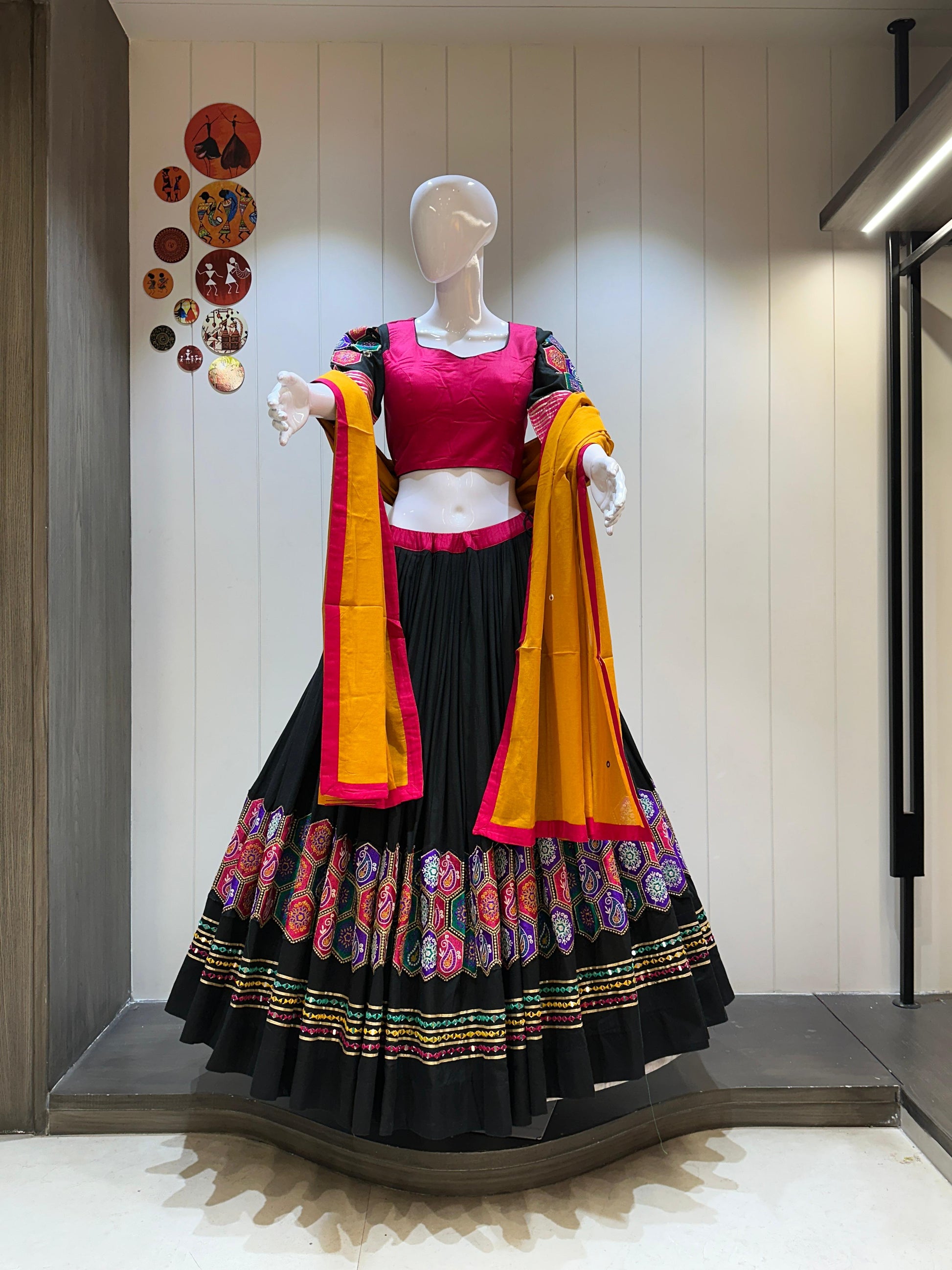 Traditional outfit with a pink blouse, black skirt, and orange dupatta on a mannequin against a neutral wall.