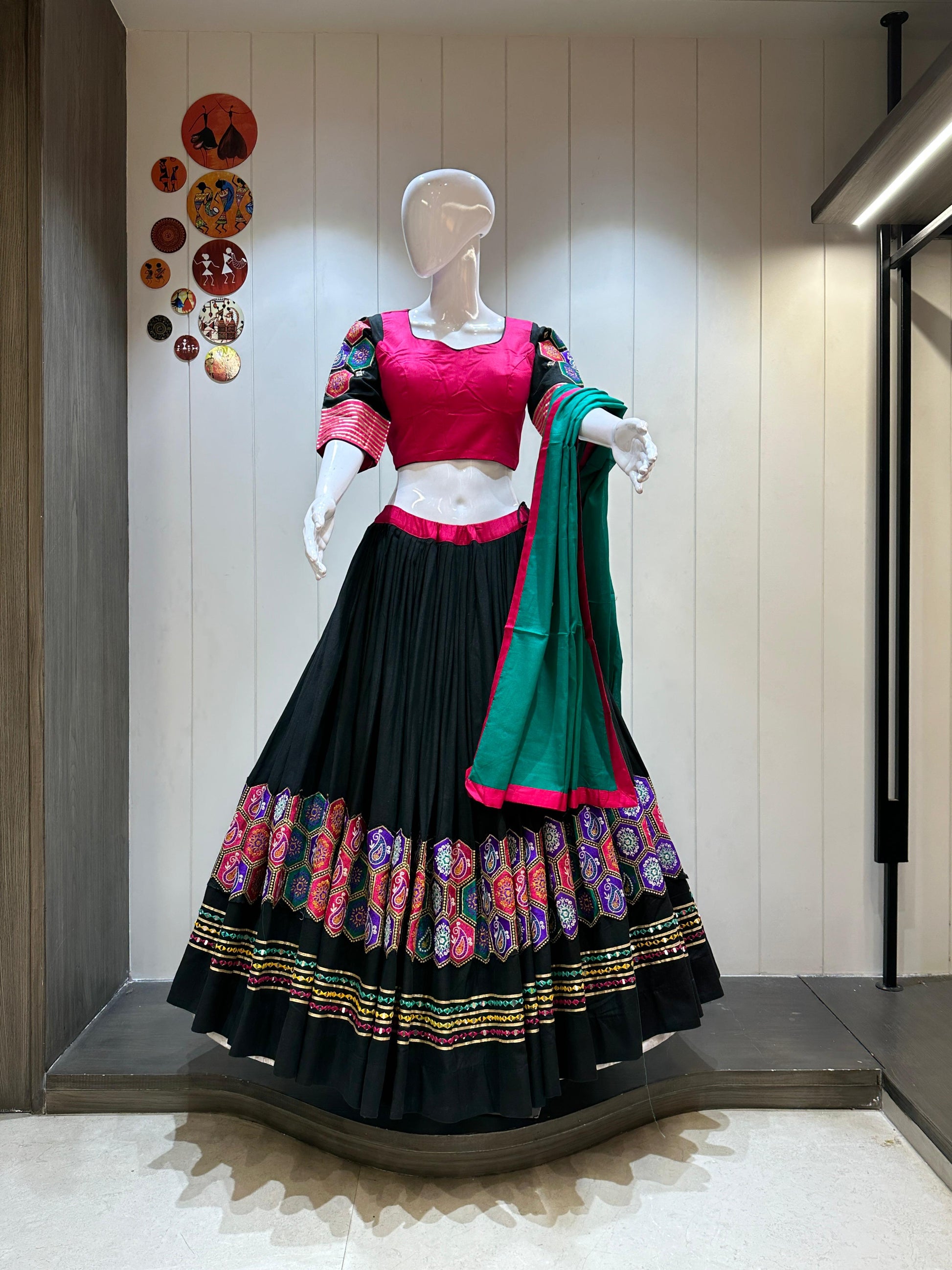 Mannequin wearing a traditional outfit with a pink blouse and black skirt with colorful patterns, against a neutral wall.