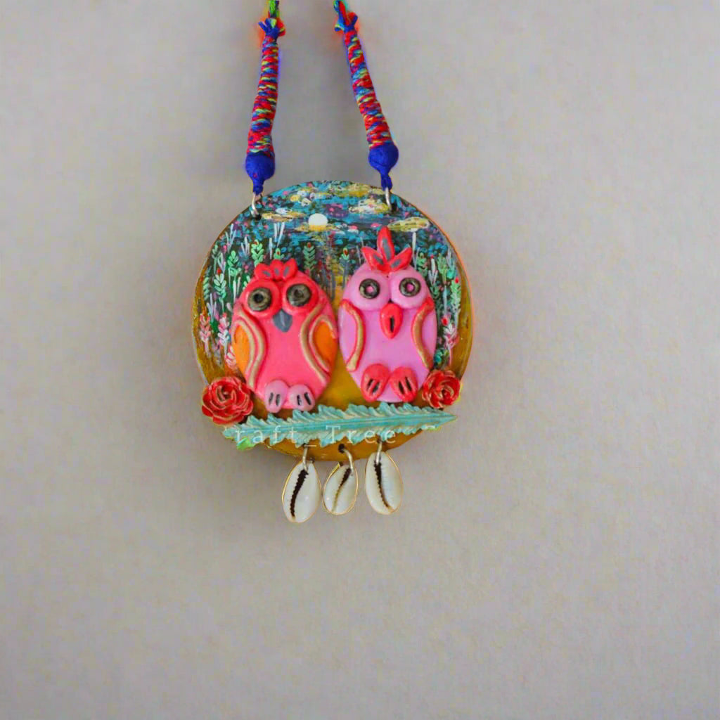 A handcrafted fabric beads jewellery featuring two owl designs in pink and blue on a light fabric background with floral accents and a red bead.